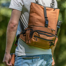 Load image into Gallery viewer, A person outdoors wearing a brown canvas backpack with black straps and multiple zippered compartments, shown from the back.