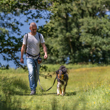 Load image into Gallery viewer, A man walks a dog on a leash through a grassy field with trees in the background. The man wears a white t-shirt, blue jeans, and a black dog harness with a small bag, while the dog wears a black leash.