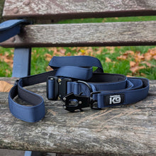Load image into Gallery viewer, A navy blue nylon dog leash with black hardware and a black brand patch reading 'TACTICAL K9' is coiled on a weathered wooden bench outdoors with grass and fallen leaves in the background.