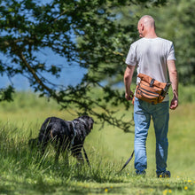 Load image into Gallery viewer, A man seen from behind walking in a grassy field with a black dog on a leash, wearing a white t-shirt, blue jeans, and a brown crossbody bag.
