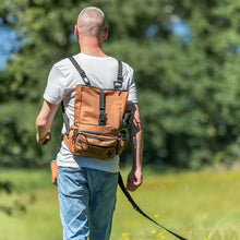 Load image into Gallery viewer, A man viewed from behind walking outdoors in a grassy field, wearing a brown canvas backpack with black straps and buckles over a white t-shirt and blue jeans.

