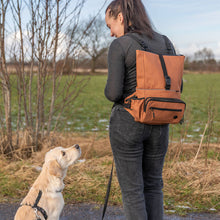 Load image into Gallery viewer, A woman wearing a tan dog backpack stands outdoors on a path with her golden retriever dog looking up at her. The backpack is a warm tan/caramel color with black straps and zipper details.
