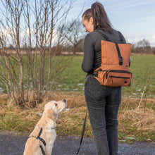 Load image into Gallery viewer, A woman wearing a brown dog backpack stands outdoors on a path with her golden retriever dog, showing how the backpack fits and looks when worn. The background features a grassy field and bare winter trees.
