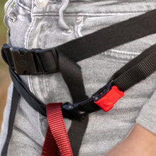 Load image into Gallery viewer, Close-up of a black webbing harness with red and black straps, black plastic quick-release buckles, and a red release button, worn around the waist over gray denim jeans.
