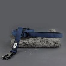 Load image into Gallery viewer, A blue webbed dog leash with a black buckle and a black logo patch, displayed on a textured gray stone prop against a dark gray studio background.
