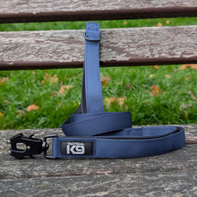 Load image into Gallery viewer, A blue tactical dog leash with a black metal quick-release buckle and a black brand patch reading 'TACTICAL K9', resting on a weathered wooden bench with green grass blurred in the background.
