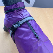 Load image into Gallery viewer, A purple dog raincoat with black and gray straps, buckles, and a front zipper, shown fitted on a dog sitting on a light wood table.
